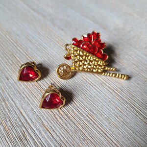 May include: A set of two red heart-shaped earrings and a gold-tone brooch shaped like a wheelbarrow filled with red hearts.