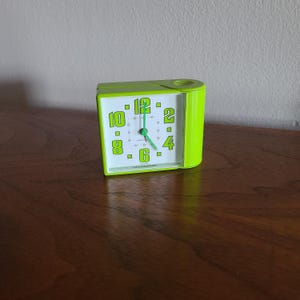 May include: A lime green rectangular alarm clock with a white face, featuring bold lime green numerals for 12, 2, 4, 6, 8, and 10. The clock has thin green hour, minute, and second hands, with a circular indentation on the top right corner of the casing.