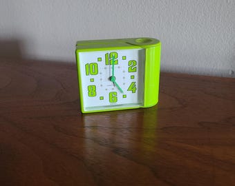 Könnte beinhalten: Ein limettengrüner, rechteckiger Wecker mit weißem Zifferblatt und fetten limettengrünen Ziffern für 12, 2, 4, 6, 8 und 10. Die Uhr hat dünne grüne Zeiger für Stunden, Minuten und Sekunden sowie eine kreisförmige Vertiefung an der oberen rechten Ecke.