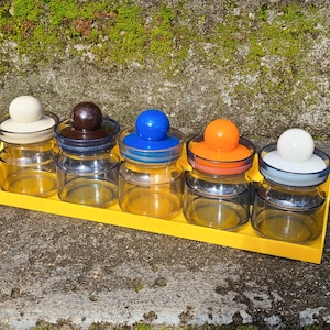 May include: A set of five clear glass jars with colorful lids in white, brown, blue, orange, and white, arranged on a bright yellow tray. The jars are small and round, perfect for storing spices or small items.