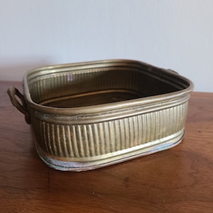 May include: A brass planter with two handles. The planter has a ribbed design and a square shape.