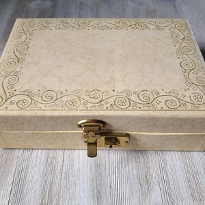 May include: A cream-colored, rectangular box with a decorative floral design around the edge. The box has a brass latch and lock.