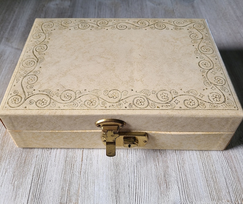 May include: A cream-colored, rectangular box with a decorative floral design around the edge. The box has a brass latch and lock.