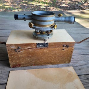May include: A vintage Bostrom surveying instrument with a grey and brass finish, featuring a central circular housing, a horizontal telescope, and adjustment knobs. It rests on a rectangular wooden storage box with metal latches and a nameplate.