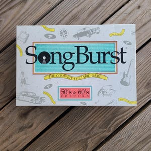 May include: A white board game box with the title "SongBurst" in large black letters. The box features illustrations of a record, car, guitar, and other 1950s and 1960s themed graphics. The game is the "50's & 60's Edition".