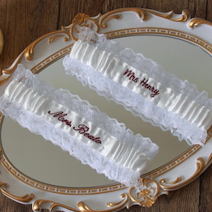 May include: Two white satin wedding garters featuring delicate lace trim and gathered fabric. Each garter is embroidered with dark red text, one reading Mrs. Henry and the other reading Mrs. Beato, displayed on an ornate oval mirror tray.