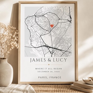Op de afbeelding: Een ingelijste kaartprint met een zwart-wit stratenplan van Parijs, Frankrijk, met een rood hartje op een specifieke locatie. De tekst luidt: JAMES & LUCY, WHERE IT ALL BEGAN, DECEMBER 16, 2020, PARIS, FRANCE, in een schreeflettertype.
