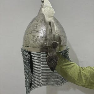 Peut inclure: Un casque en métal argenté de forme bombée et pointue, orné de motifs floraux et de volutes gravés sur toute la surface. Une protection nasale verticale en métal s'étend sur le devant et un camail en mailles est fixé à la base.