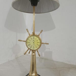 Può includere: Una lampada da tavolo in ottone con un design a timone di nave e un orologio al centro. L'orologio presenta numeri romani e la scritta "J. P. LONDON". La lampada include un alto supporto in ottone, una base circolare in legno scuro e un paralume grigio.