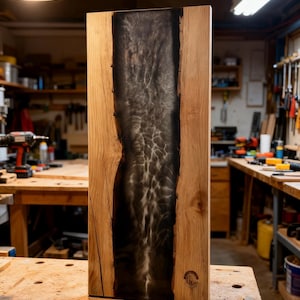 May include: A rectangular wooden board featuring a central vertical inlay of dark, translucent epoxy resin with a swirling, smoke-like pattern. The natural light-colored wood grain frames the resin, and a small circular logo is stamped on the bottom right.