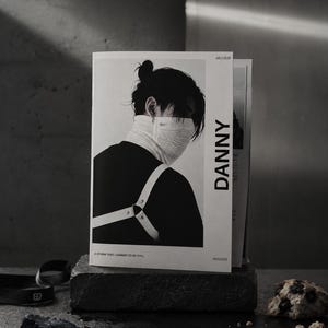 Könnte beinhalten: Ein schwarz-weißes Magazin mit dem Titel DANNY, datiert Juli 2026, das ein Porträt einer Person mit Dutt-Frisur zeigt, die eine Gesichtsmaske und ein Geschirr über einem dunklen Oberteil trägt. Das Cover enthält den Text A STORM THAT LEARNED TO BE STILL.