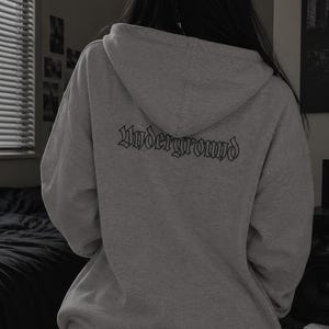 Könnte beinhalten: Ein hellgrauer Kapuzenpullover mit langen Ärmeln und einer Kapuze. Auf der Rückseite des Kleidungsstücks ist das Wort Underground in einer schwarzen, stilisierten gotischen Schrift im oberen Bereich unter der Kapuzennaht gedruckt.