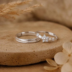 May include: Two silver-toned rings with a polished finish. One ring features a band with small engraved star details. The second ring displays a cluster of star-shaped settings, each containing a clear, sparkling, round-cut gemstone.