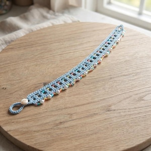 May include: A light blue crocheted bracelet featuring a repeating openwork pattern. The edges are embellished with a variety of small, colorful glass beads and white faux pearls. One end of the bracelet terminates in a single crocheted loop.
