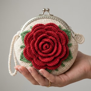 May include: A round, cream-colored crochet coin purse featuring a large, three-dimensional red rose with green leaves centered on the front. The purse has a silver-toned metal kiss-lock clasp closure and a matching cream-colored braided wrist strap.