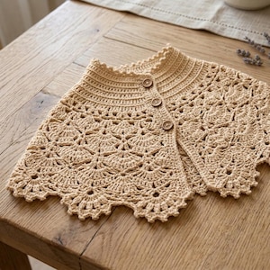 May include: A beige crochet capelet featuring a scalloped edge and a delicate fan-like pattern throughout the body. The garment has a ribbed collar and is fastened down the front with three small, round, brown wooden buttons.