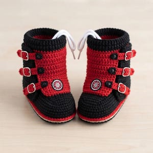 Könnte beinhalten: Ein Paar gehäkelte Stiefeletten in Schwarz und Rot mit drei horizontalen Riemen und silberfarbenen Metallschnallen an jeder Seite. Die Vorderseite jedes Schuhs zeigt ein kreisförmiges Emblem mit einem weißen Radmotiv und weiße Schnürsenkel.