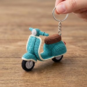 Könnte beinhalten: Ein türkisfarbener Häkel-Roller-Schlüsselanhänger mit braunem Sitz, weißem Rand und schwarzen Rädern mit grauen Zentren. Das Miniaturfahrzeug ist mit einer kurzen Kette an einem silbernen Metallring befestigt und zeigt detaillierte Garnstiche.