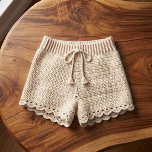 May include: Cream-colored crochet shorts featuring a high-waisted ribbed waistband with a matching drawstring tie ending in small tassels. The hem of the shorts is finished with a decorative scalloped edge pattern throughout the entire bottom.