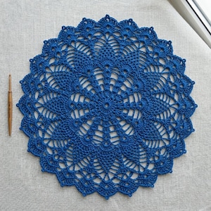 Può includere: Un centrino all'uncinetto blu di forma circolare con un intricato motivo ispirato all'ananas e un bordo smerlato. Un uncinetto in legno è posizionato a sinistra del centrino, con la misura di 2,5 mm stampata sul manico dell'attrezzo.