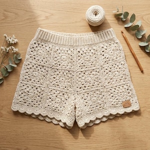May include: Off-white crochet shorts featuring a square motif pattern with a scalloped hem and a high-waisted ribbed waistband. A small rectangular brown leather-like tag with a mountain icon is attached to the lower right corner of the shorts.