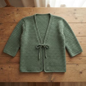 May include: A sage green crochet cardigan featuring a V-neckline, three-quarter length sleeves, and a thin tie closure at the center front. The garment is constructed with a consistent horizontal stitch pattern throughout the entire piece.