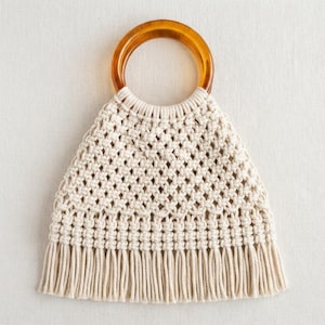 Puede incluir: Un bolso de macramé de color crema con un asa circular de resina en tono ámbar. El cuerpo del bolso está elaborado con un patrón repetitivo de rombos anudados y rematado con un fleco largo y recto a lo largo del borde inferior de la estructura.