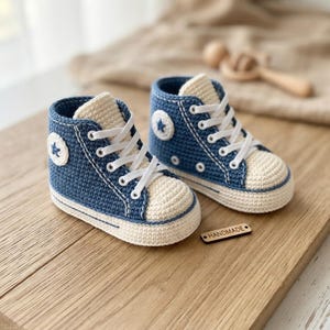 Op de afbeelding: Een paar blauwe en crèmekleurige gehaakte hoge sneakers met witte veters, voorzien van een ronde patch met een blauwe ster aan de zijkant, een crèmekleurige neus en een klein houten label met het woord HANDMADE in zwarte letters.