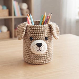 Könnte beinhalten: Ein zylindrischer Stiftehalter aus beigem Häkelgarn in Hundeform mit Schlappohren, schwarzen Perlenaugen und einer weißen Schnauze mit aufgestickter schwarzer Nase und Mund. Der Halter ist mit verschiedenen Buntstiften und Markern gefüllt.