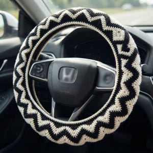 May include: A crochet steering wheel cover featuring a repeating black and cream zigzag pattern. The cover is fitted around a black steering wheel with a central Honda logo, showcasing a textured yarn construction with a consistent chevron design.