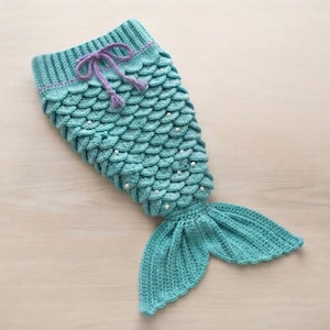 May include: A turquoise crochet mermaid tail blanket featuring a ribbed top edge with a purple drawstring tie. The body is constructed with overlapping scale-like stitches accented by small white beads, ending in a wide, ribbed fin at the base.