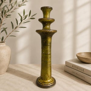 Puede incluir: Un candelabro alto de color verde oliva con un acabado texturizado y desgastado. El objeto presenta un diseño escalonado de varios niveles con una base ancha, una columna central cilíndrica y dos plataformas circulares que llevan a la copa superior.
