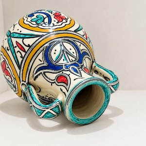 Puede incluir: Una jarra de cerámica con base color crema que presenta patrones pintados a mano en tonos vibrantes de turquesa, azul intenso, amarillo y rojo. El recipiente tiene un cuerpo redondeado, cuello ancho y dos asas simétricas con motivos geométricos.