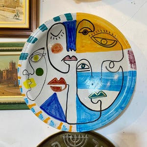 Puede incluir: Un plato decorativo redondo de cerámica con un diseño abstracto pintado a mano de tres rostros. La obra utiliza contornos negros marcados con bloques vibrantes de pintura amarilla, azul y verde. Los rostros están estilizados.