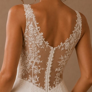 Puede incluir: Un vestido blanco sin mangas con un panel trasero de ilusión transparente adornado con intrincados bordados de encaje floral blanco. El corpiño incluye una fila vertical de pequeños botones cubiertos de tela en la espalda, sobre una falda lisa.