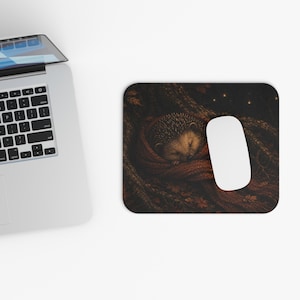 May include: A rectangular mouse pad featuring a graphic illustration of a small hedgehog curled up and sleeping inside a textured, dark orange knitted scarf, set against a dark, forest-themed background with scattered autumn leaves and soft light.