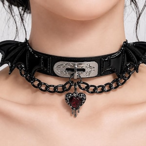 May include: A black choker featuring glossy bat wings on each side, a central silver-toned metal plate with an engraved pattern, a black chain draped across the front, and a dangling dark red heart-shaped pendant with a metallic drip design.