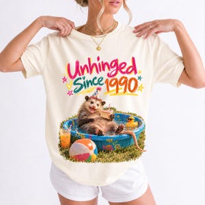 Opossum in party hat in a kiddie pool with cake, 90s film photo style, text "Unhinged Since 1990"
