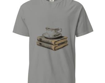 May include: A grey short-sleeve t-shirt featuring a graphic print of a white teacup on a saucer resting atop a stack of two vintage-style books with tan pages and dark covers. The design includes faint wisps of steam rising from the teacup.