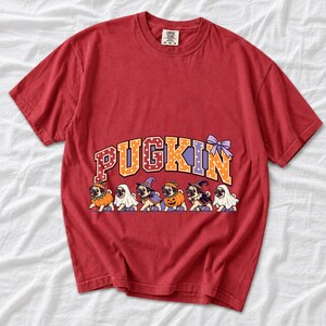 Six pugs in pumpkin, ghost and witch costumes under the arched word PUGKIN, lavender and orange polka-dot varsity style