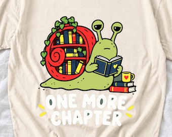 Puede incluir: Una camiseta color crema con un gráfico de un caracol verde claro con una concha roja en forma de estantería llena de libros. El caracol está leyendo un libro, con una taza amarilla y dos libros cerca. El texto dice: ONE MORE CHAPTER.