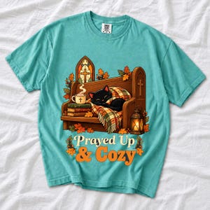 Black cat curled on a church pew with blanket and cocoa among autumn leaves, text "Prayed Up & Cozy", retro illustration
