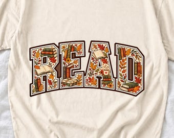 Puede incluir: Una camiseta de color crema con la palabra READ en letras grandes de bloque. Las letras están rellenas con ilustraciones de libros apilados, libros abiertos, una taza humeante con un corazón y hojas de otoño en tonos naranja, rojo y marrón.