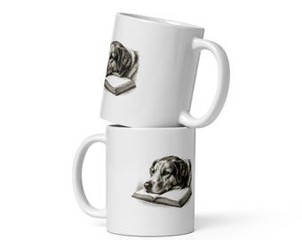 May include: Two white ceramic mugs stacked vertically. Each mug features a black and white detailed illustration of a dog resting its head on an open book. The mugs have a smooth, glossy finish with a standard handle on the side of each vessel.