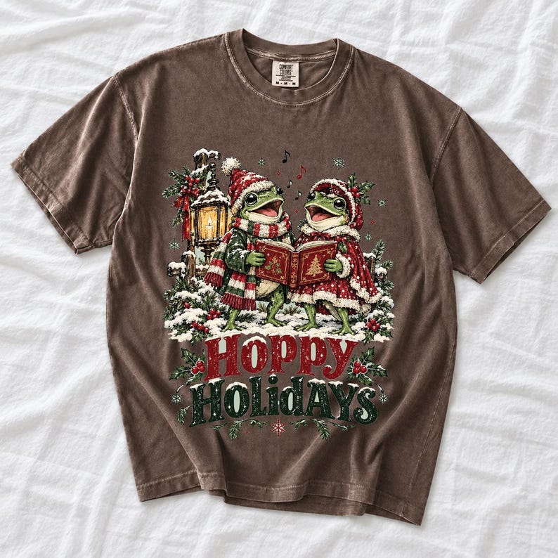 Two frogs singing carols in the snow with a lantern and songbook, text Hoppy Holidays