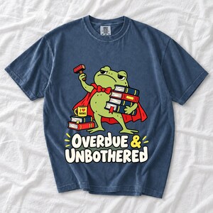 Grumpy cartoon frog librarian holding a stamp and a stack of books with the text Overdue & Unbothered