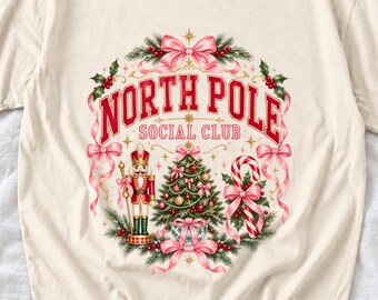 May include: A cream-coloured t-shirt featuring a graphic print of a Christmas wreath with the text "NORTH POLE SOCIAL CLUB". The design includes a nutcracker, a decorated pine tree, a candy cane, holly leaves, and pink ribbons.