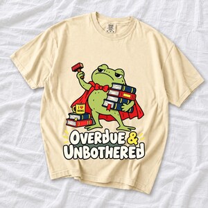 Grumpy cartoon frog librarian holding a stamp and a stack of books with the text Overdue & Unbothered