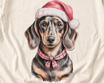 May include: A cream-colored t-shirt featuring a graphic illustration of a black and tan dachshund wearing a red and white Santa hat and a red and white plaid scarf around its neck, centered on the fabric.