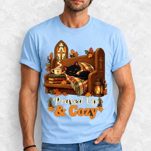 Black cat curled on a church pew with blanket and cocoa among autumn leaves, text "Prayed Up & Cozy", retro illustration